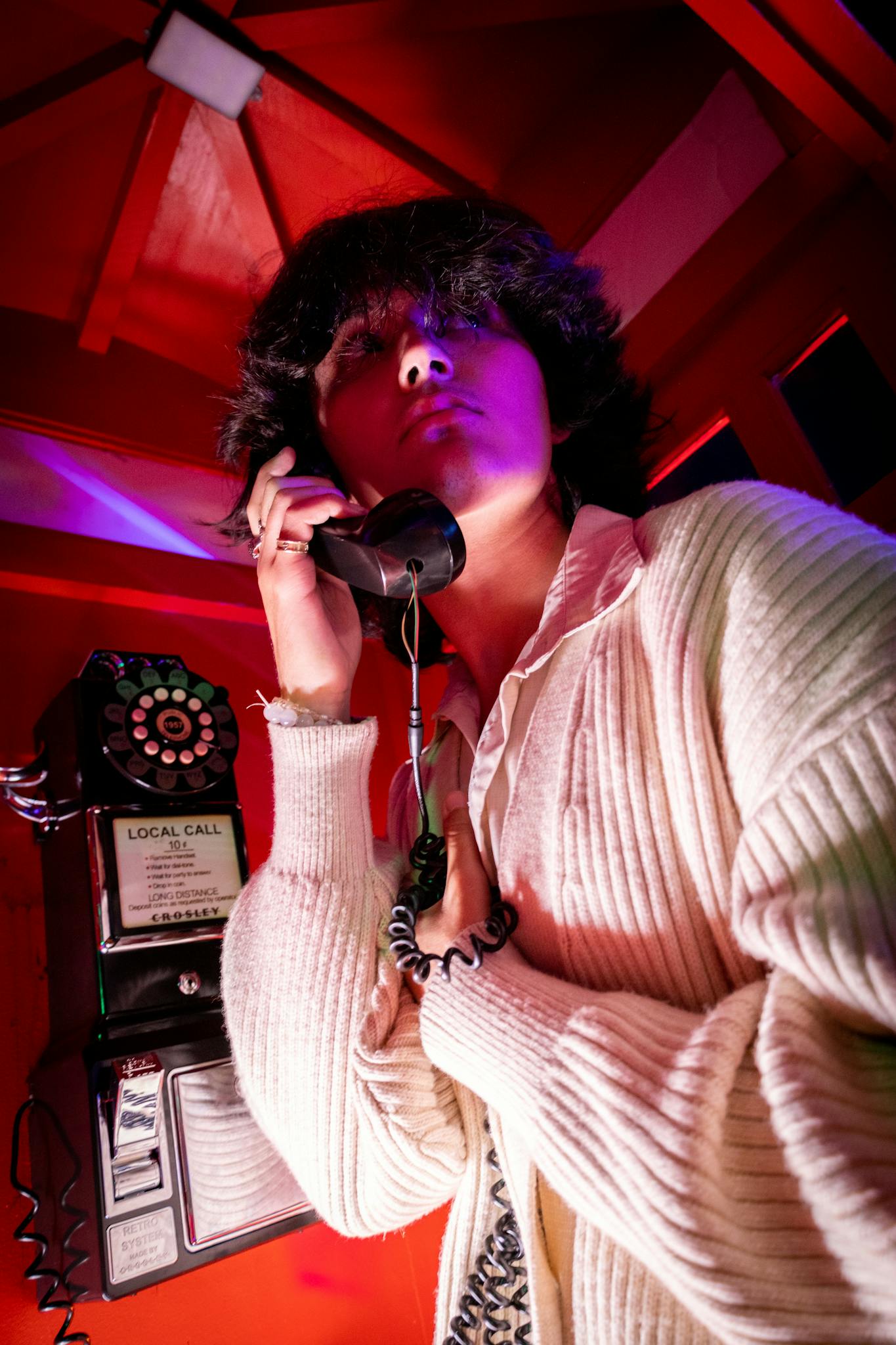 A stylish portrait of a young adult in a vintage phone booth, showcasing retro fashion and dramatic lighting.